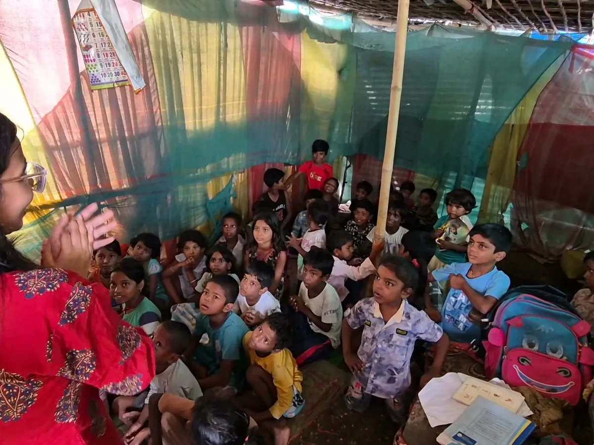 A teacher speaking to a large group of children seated on the floor of a community classroom built under patchwork tarpaulins.