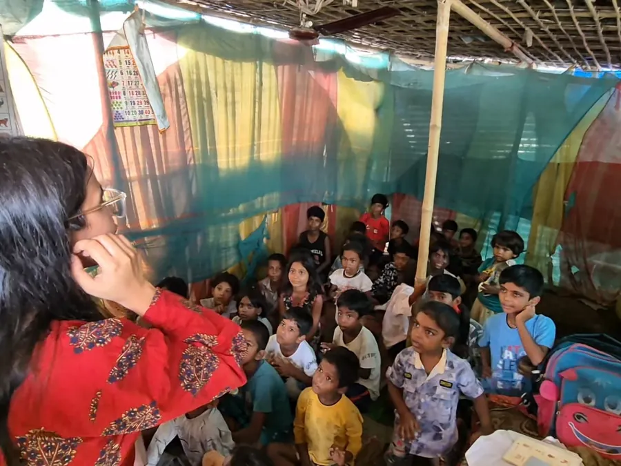 A teacher leading a session for children seated under patchwork tarpaulins.