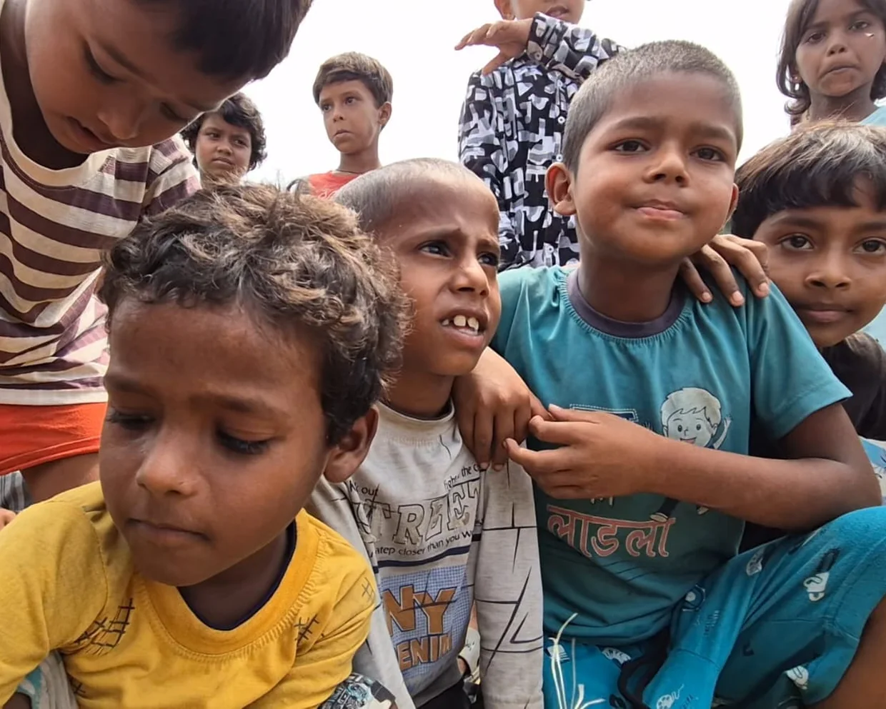 A close group of children sitting together outdoors, leaning on one another, mid-conversation.